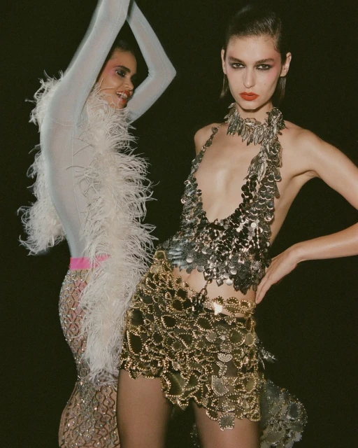 Two fashion models pose in a dark studio wearing shimmering, metallic dresses; one silver, the other gold, standing closely together.