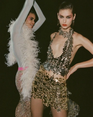 Two fashion models pose in a dark studio wearing shimmering, metallic dresses; one silver, the other gold, standing closely together.