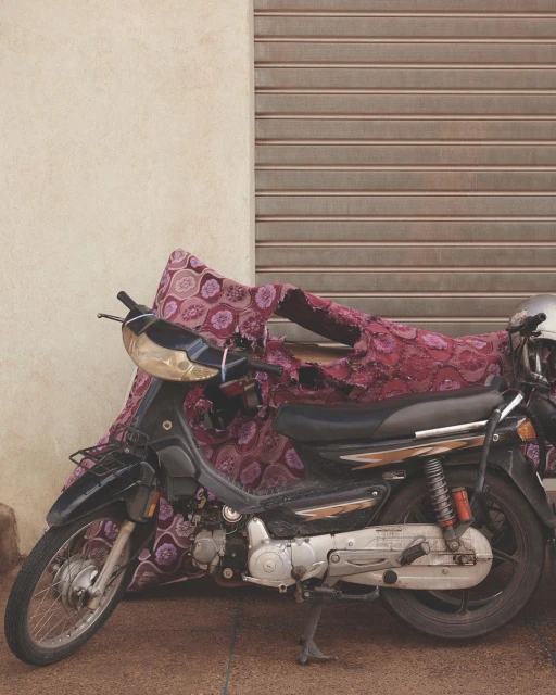 Motorcycle parked on the street with patterned textile cover, adjacent to a closed roller shutter and wall on a sunny day.