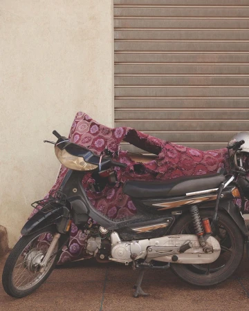 Motorcycle parked on the street with patterned textile cover, adjacent to a closed roller shutter and wall on a sunny day.