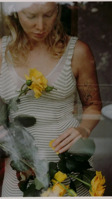 Woman in textured white dress arranges yellow roses, seen through a window with reflections of greenery and light.
