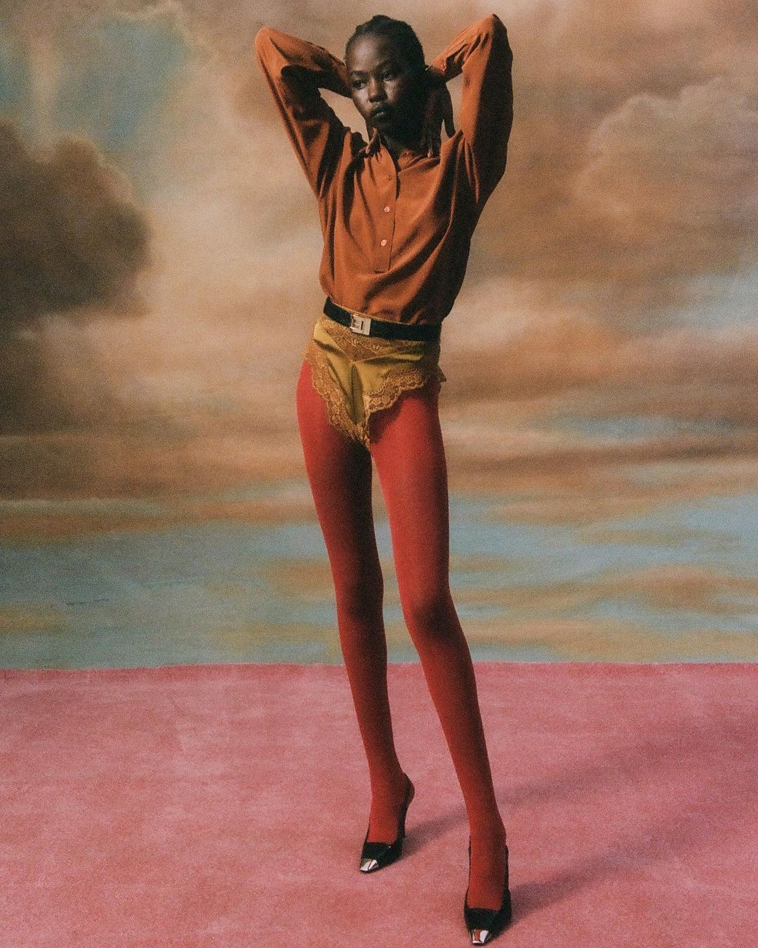 Model in rust shirt and red tights posing against a painted cloud backdrop by Hans Neumann