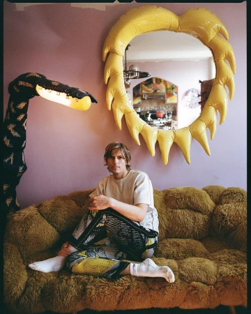 Man seated on a furry mustard couch in a pink room featuring a yellow inflatable sun mirror and a tall sculptural lamp