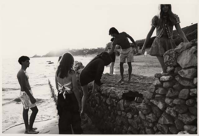 Black and white shot of a group of people on a beach near a stone wall, some standing and some climbing over it, under bright light.