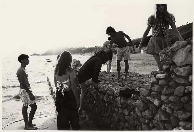 Black and white shot of a group of people on a beach near a stone wall, some standing and some climbing over it, under bright light.