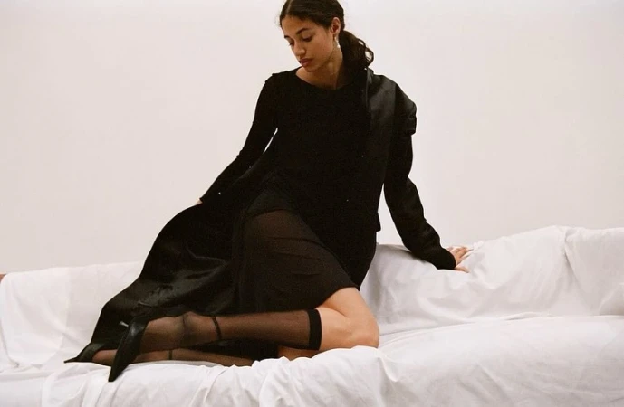 Woman in a black dress and sheer stockings seated on a white draped sofa against a plain white background