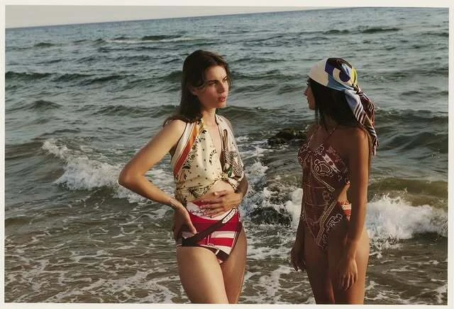 Two women in swimwear stand on a sandy beach, ocean waves behind them. One wears a headscarf in a medium outdoor shot.
