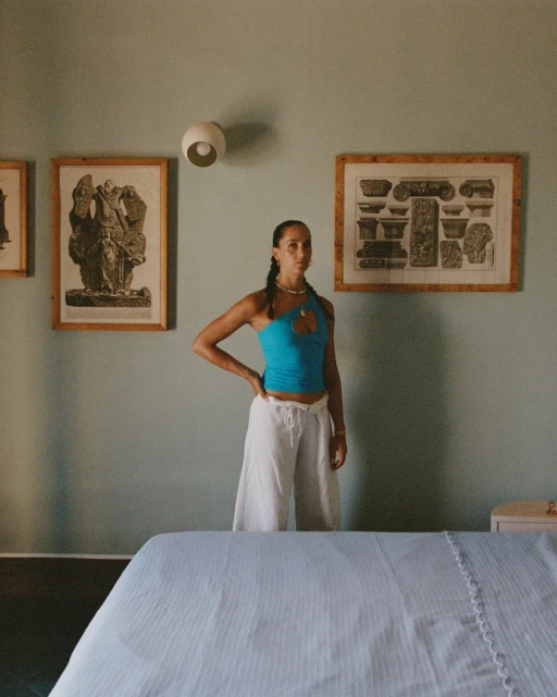 Young woman in blue top and white pants posing in an indoor setting with artwork on the wall and a bed in the foreground.