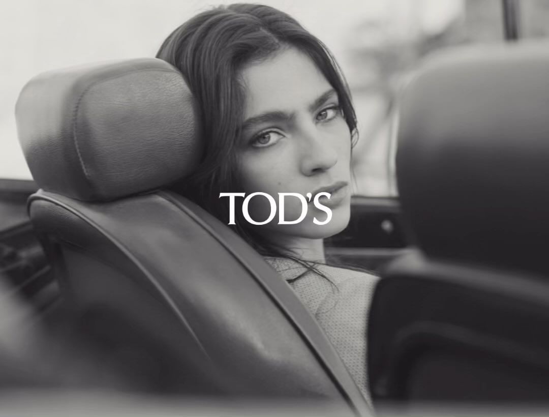 Monochrome close-up of a woman with long dark hair sitting in a vintage car; fashion shot with the logo TOD'S visible in the center.