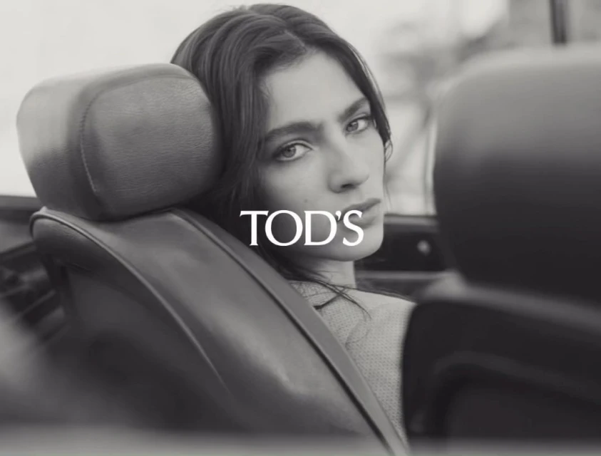 Monochrome close-up of a woman with long dark hair sitting in a vintage car; fashion shot with the logo TOD'S visible in the center.