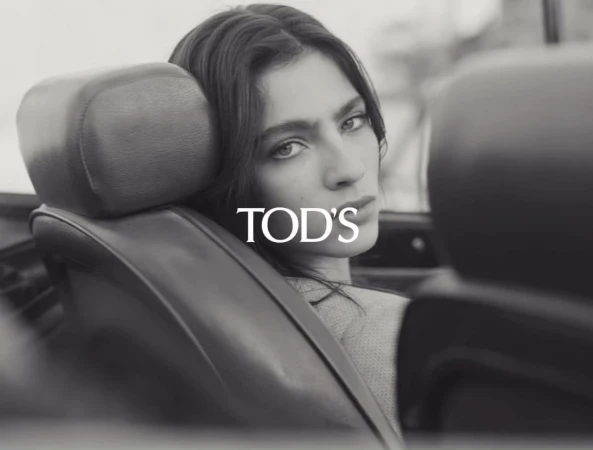 Monochrome close-up of a woman with long dark hair sitting in a vintage car; fashion shot with the logo TOD'S visible in the center.