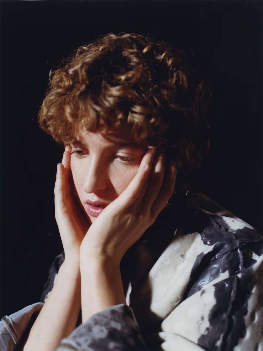 Portrait of a young woman with curly hair resting her face in her hands, with a dark background and studio lighting.