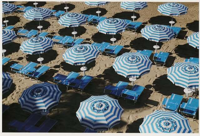 Aerial shot of a sandy beach dotted with evenly spaced blue and white striped umbrellas and matching beach chairs, creating a vacation scene.