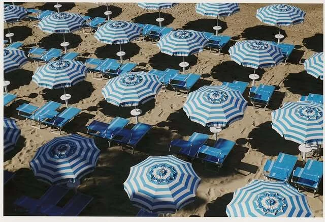 Aerial shot of a sandy beach dotted with evenly spaced blue and white striped umbrellas and matching beach chairs, creating a vacation scene.