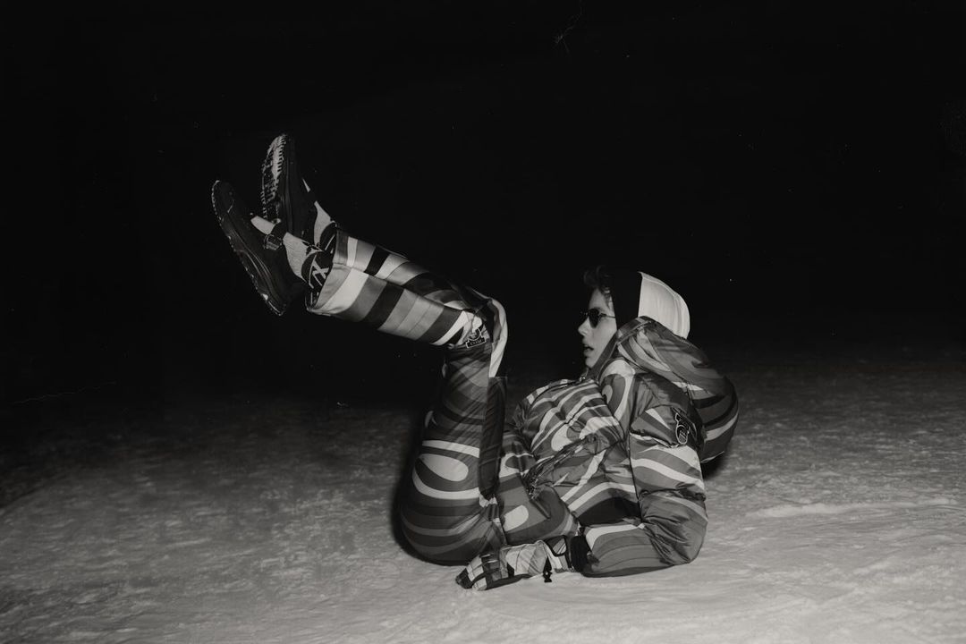 Monochrome image of a person in a striped snowsuit, sunglasses, and a cap, lying on their back in the snow with legs raised.