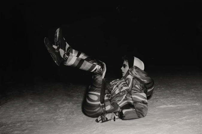 Monochrome image of a person in a striped snowsuit, sunglasses, and a cap, lying on their back in the snow with legs raised.