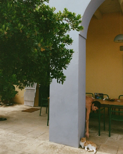 Woman interacts with a rabbit outdoors near a building with an arched doorway, enhanced by verdant trees and natural light.