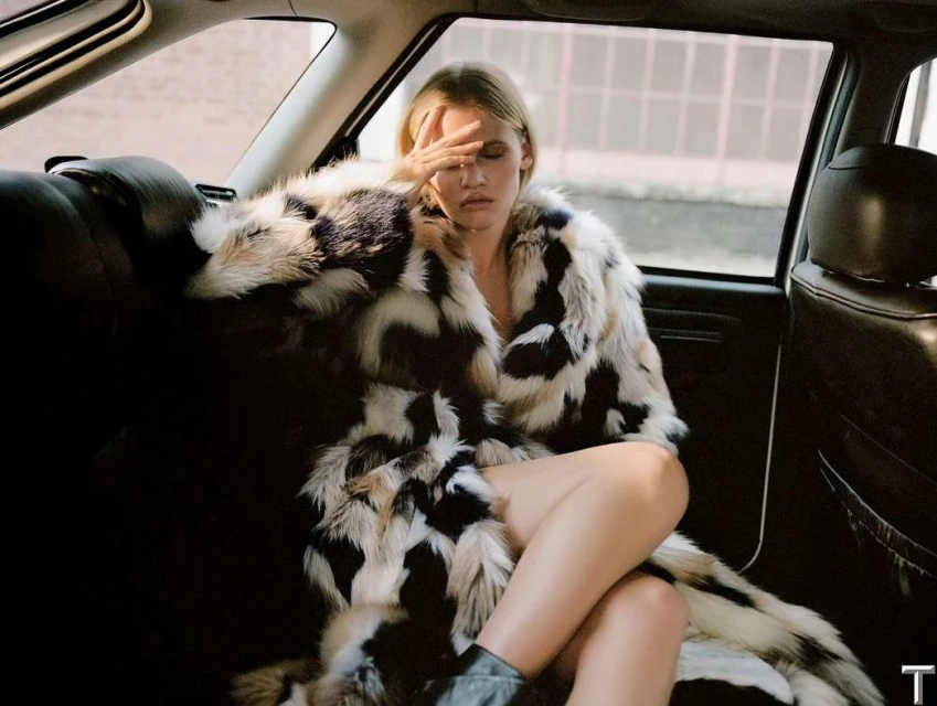 Blonde woman in a fur coat sitting in the back of a car with hand over her face looking tired or shielded from light.