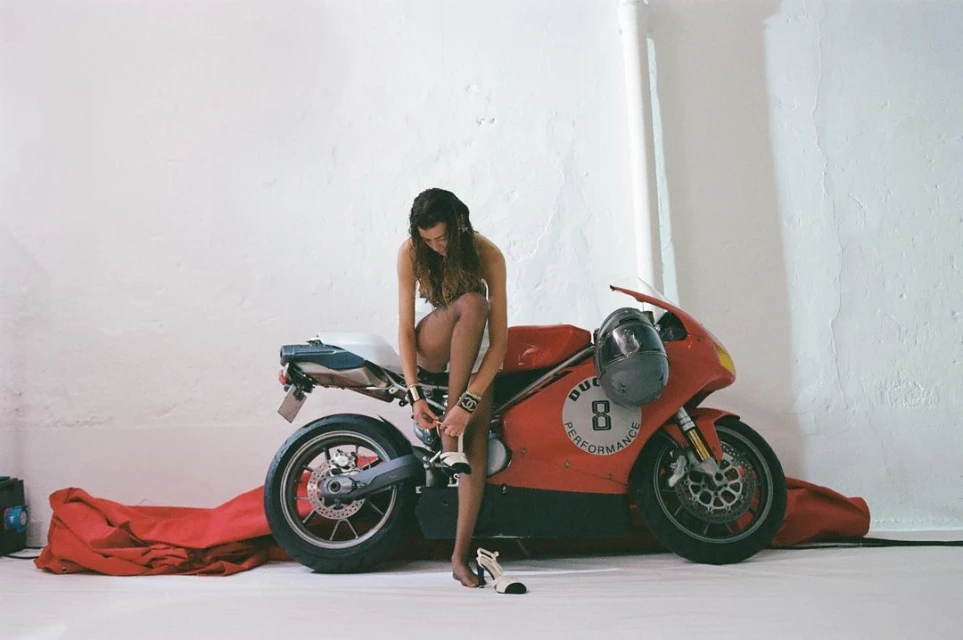 A woman in black lingerie ties her shoe while sitting on a red and white Ducati motorcycle inside a photo studio with white walls.