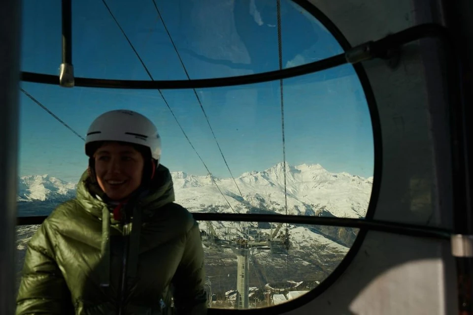 Woman in a ski gondola with helmet and green puffer jacket. A view of snow-capped mountains and ski resort infrastructure are visible through the window.