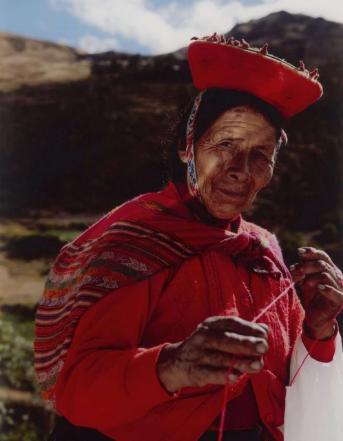 Elderly Indigenous woman in Peru weaving with bright red traditional clothing and hat in an outdoor setting with a hilly backdrop.