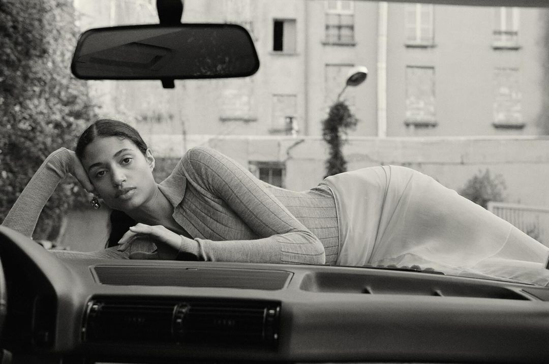 Black and white studio shot of a woman lying on the hood of a car with a building in the background, captured in a medium shot.