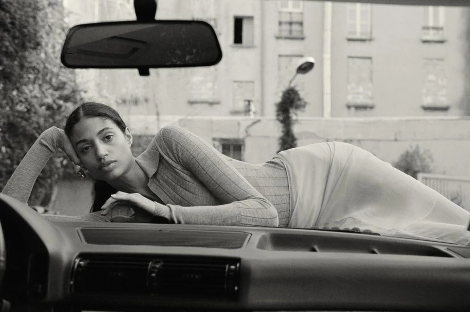 Black and white studio shot of a woman lying on the hood of a car with a building in the background, captured in a medium shot.