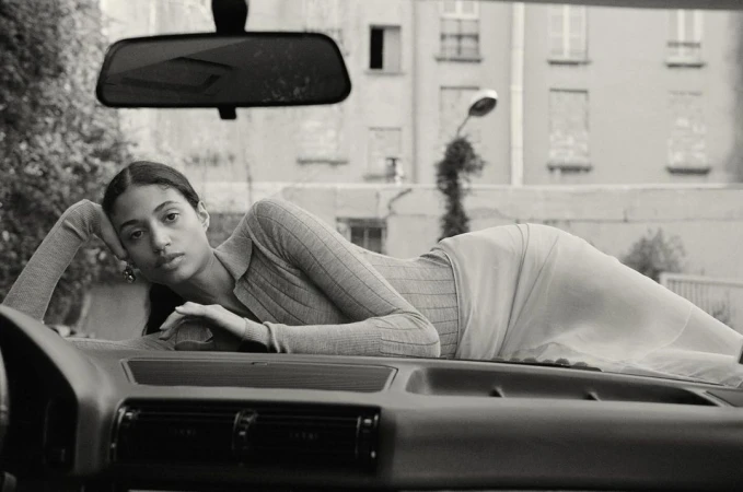Black and white studio shot of a woman lying on the hood of a car with a building in the background, captured in a medium shot.