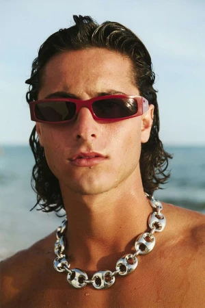 Close-up portrait of a man wearing red sunglasses and a chunky silver chain necklace against an ocean background.