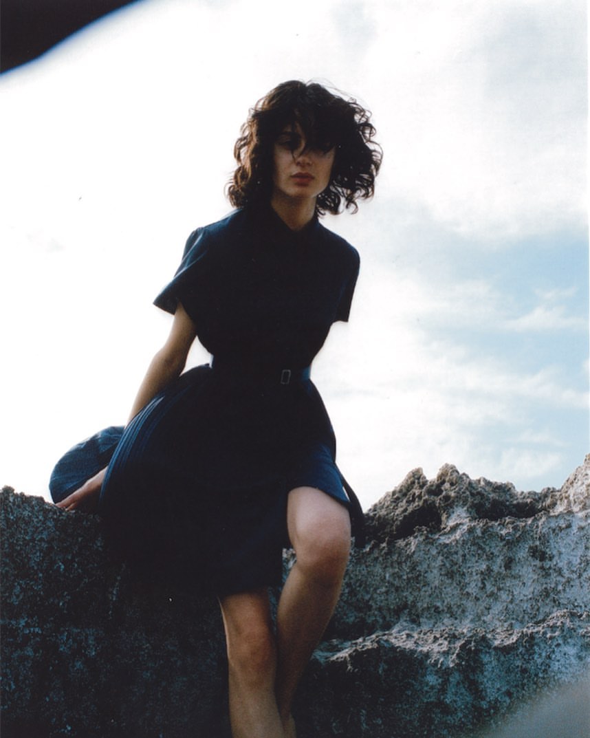 Woman with dark curly hair wearing a dark dress seated on rocky terrain under an overcast sky by Hans Neumann