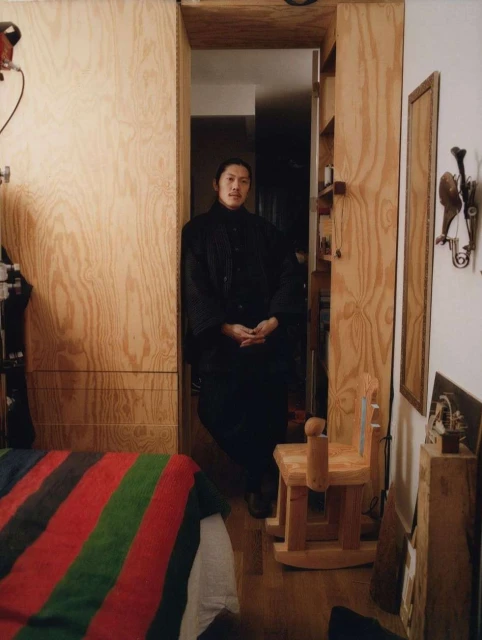 Portrait of an Asian man in a dark outfit standing in a doorway with wooden walls, complemented by a child's wooden rocking chair.