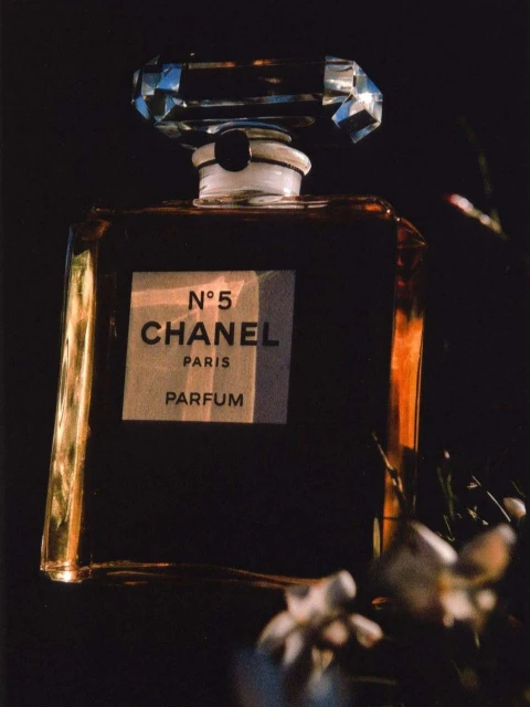 Close-up product shot of Chanel No. 5 Parfum bottle featuring a faceted crystal stopper, gold accents, and white floral details in the foreground.