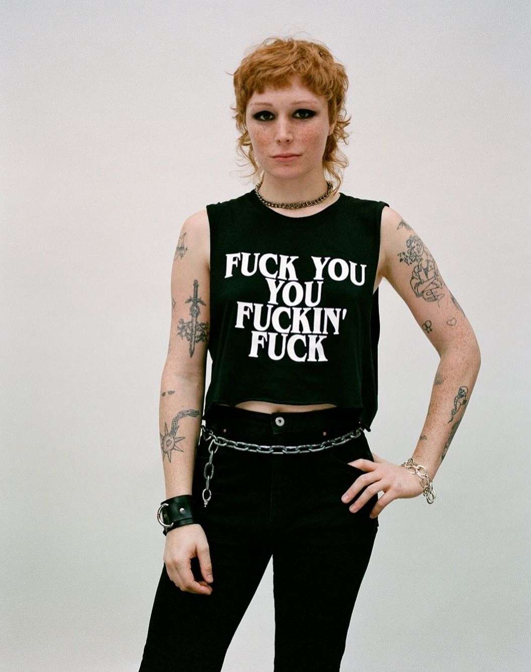 A stylish woman with a mullet poses in a studio wearing a black crop top with white text, dark pants, and chain accessories. 