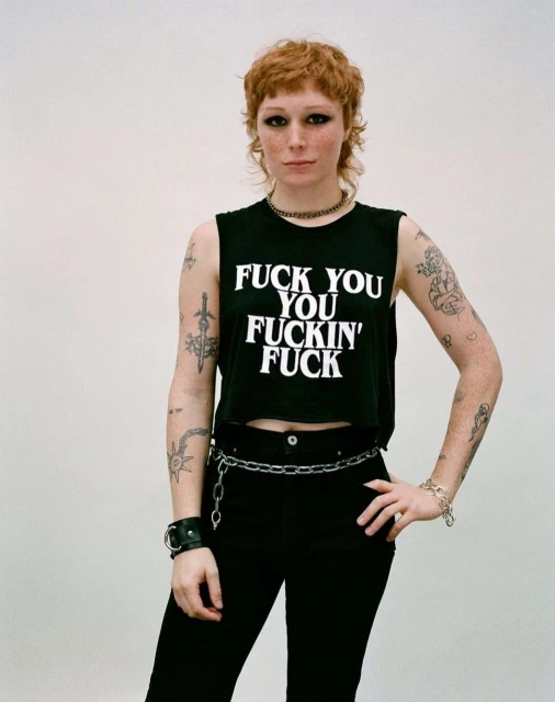 A stylish woman with a mullet poses in a studio wearing a black crop top with white text, dark pants, and chain accessories. 