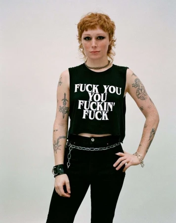 A stylish woman with a mullet poses in a studio wearing a black crop top with white text, dark pants, and chain accessories. 