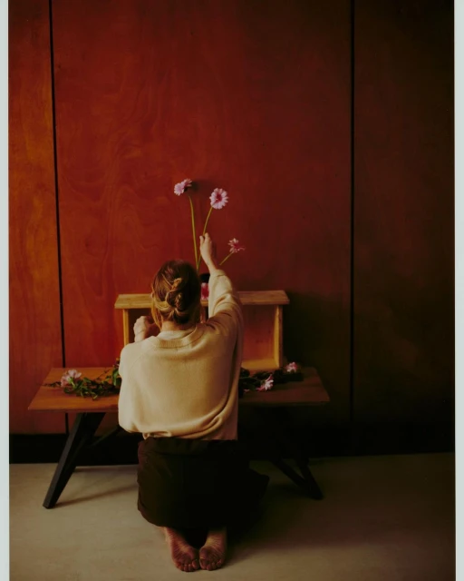 Back view of adult female arranging gerbera daisies on a wood bench in front of warm-toned wall in a minimalist indoor space.