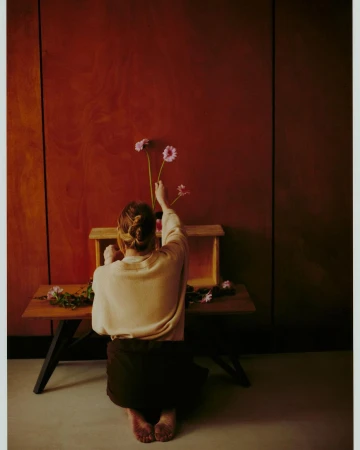 Back view of adult female arranging gerbera daisies on a wood bench in front of warm-toned wall in a minimalist indoor space.