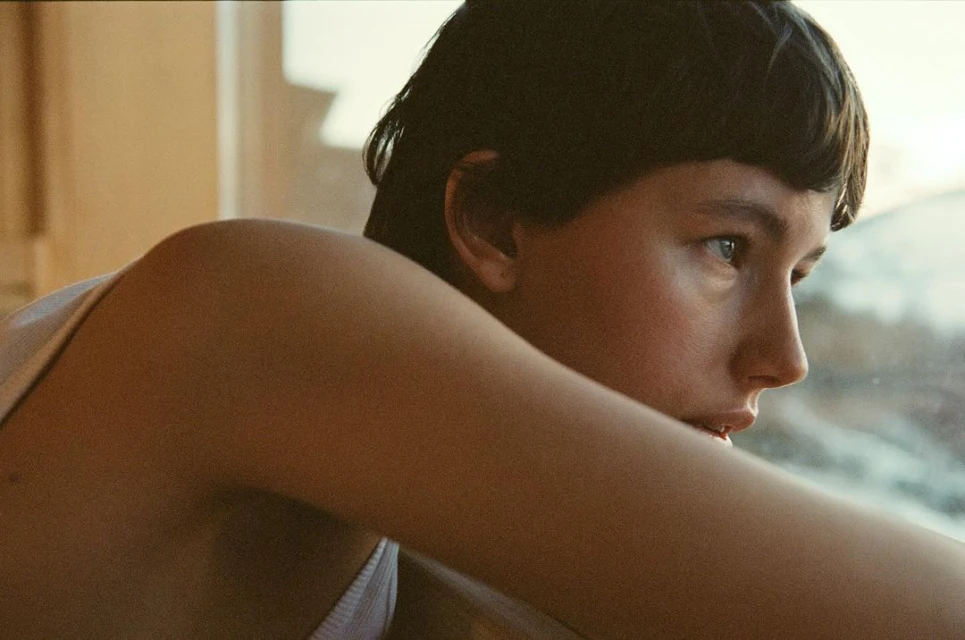 Close-up of a young person with short dark hair and fair skin looking out a window in warm indoor light.