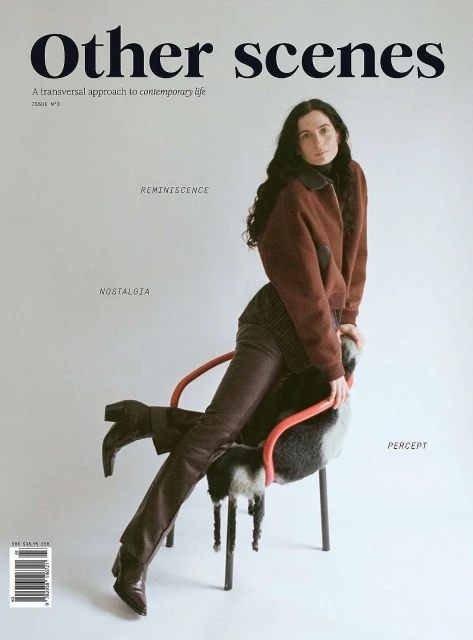 Magazine cover featuring a woman seated on a chair wearing a brown jacket and pants, photographed indoors against a grey backdrop.