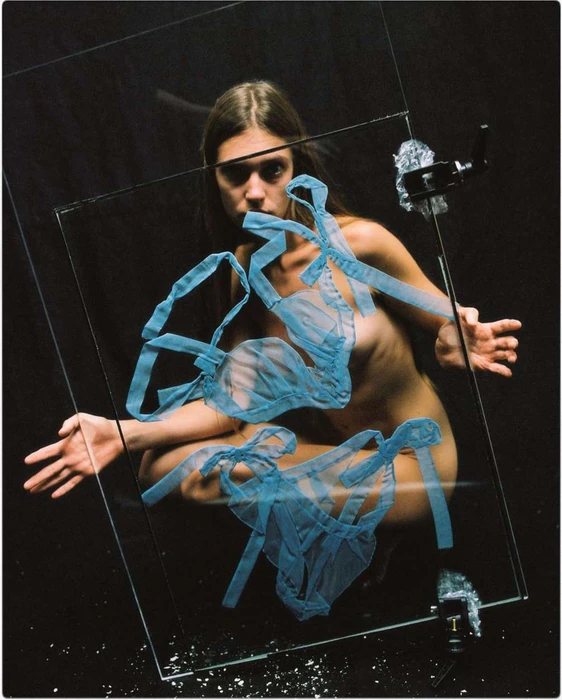 Woman crouching behind a tilted pane of glass with blue mesh lingerie taped to it against a dark background by Hans Neumann