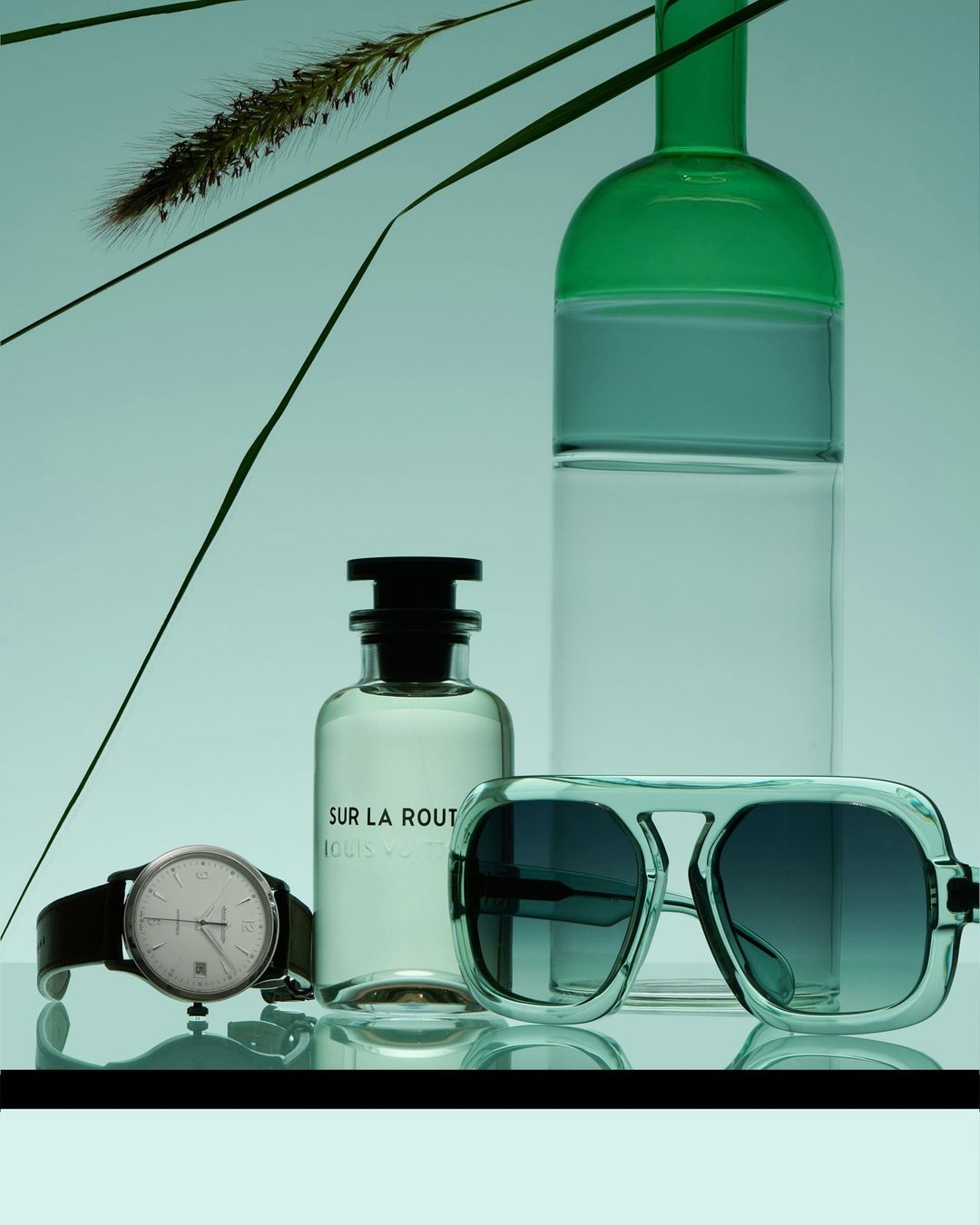 Still life photo of sunglasses, a wristwatch, and a Louis Vuitton perfume bottle against a light blue background with grass elements.