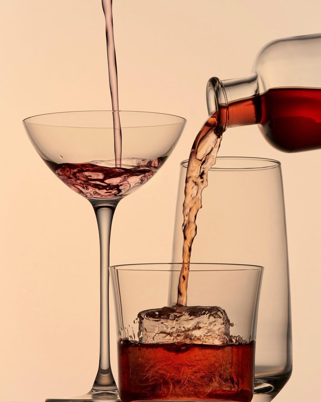 Liquid being poured into various glassware including a coupe glass, a lowball glass with ice, and a highball glass on a beige background.