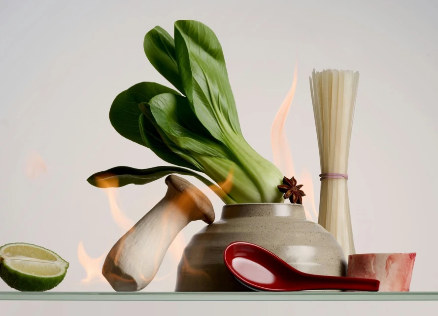 Food photography: Bok choy and mushroom in a bowl with noodles, star anise, and lime on a reflective surface against a white backdrop.