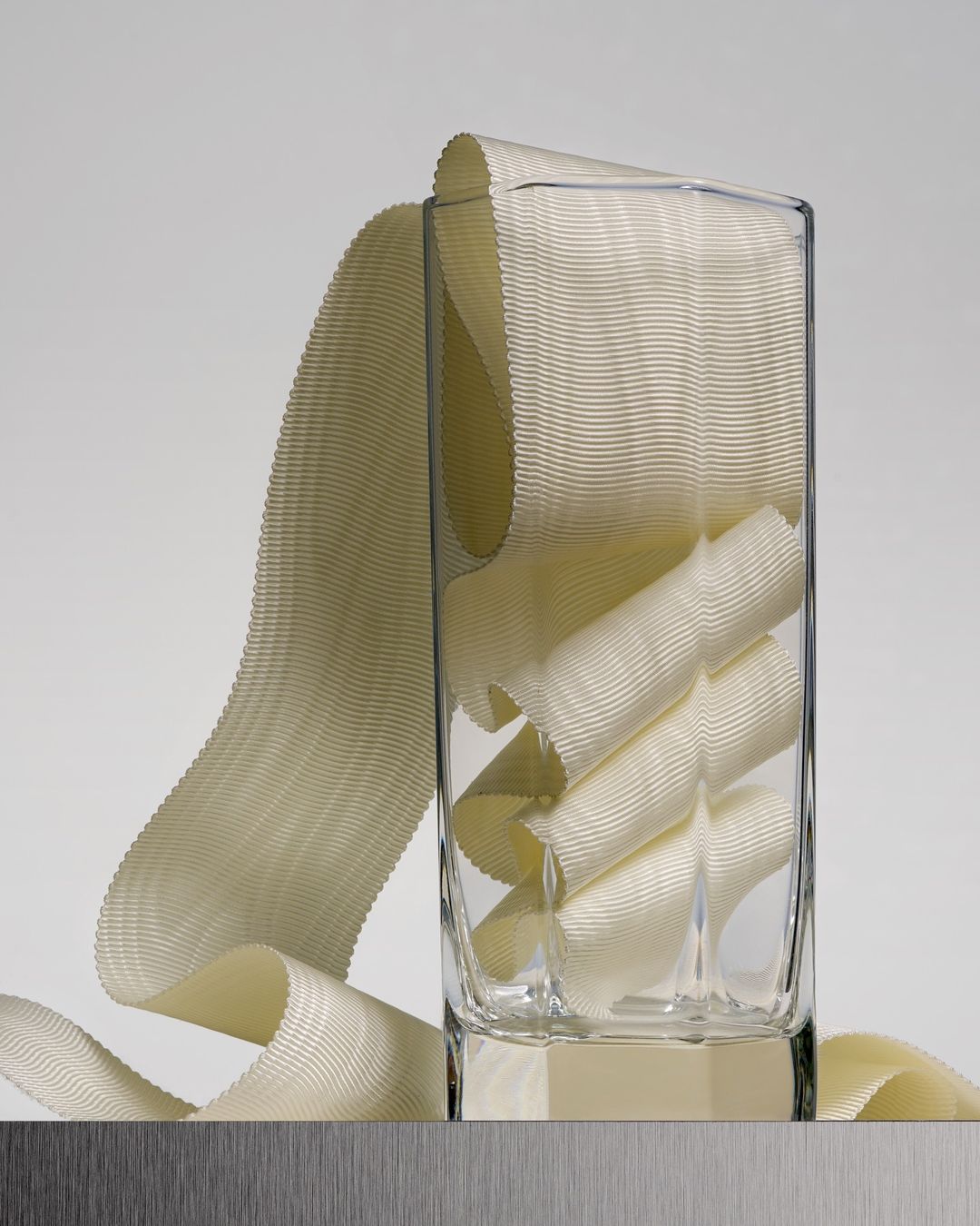 Textured ivory ribbon cascading and folded inside a clear square glass on a brushed metal surface against a light grey background.