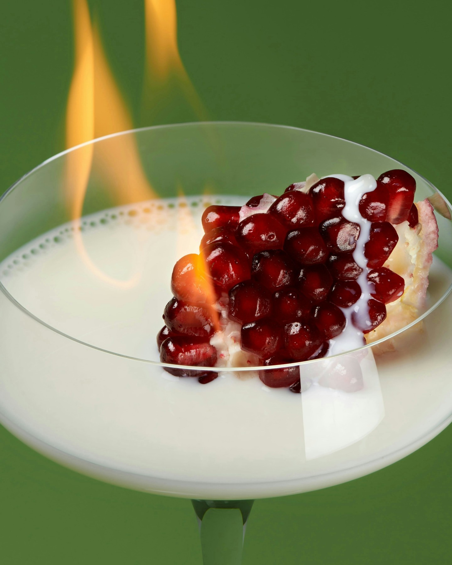 Close-up of a cocktail glass with a pomegranate wedge on fire, sitting in a white liquid, set against a solid green backdrop.