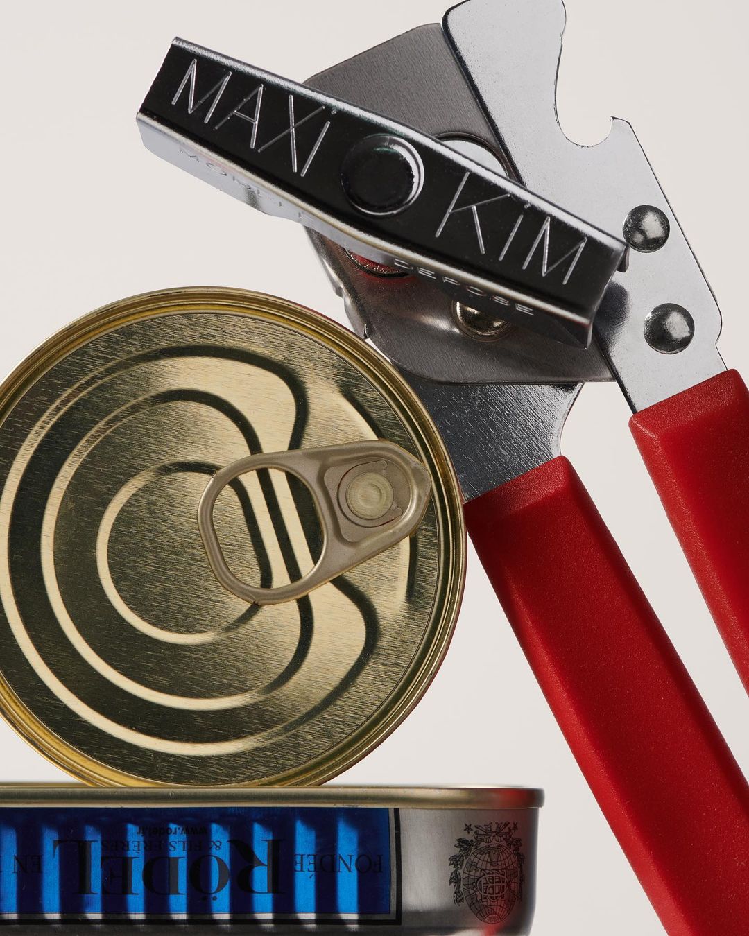 Close-up of a can opener cutting open a golden tin can, with another sealed can visible beneath, all against a white backdrop.