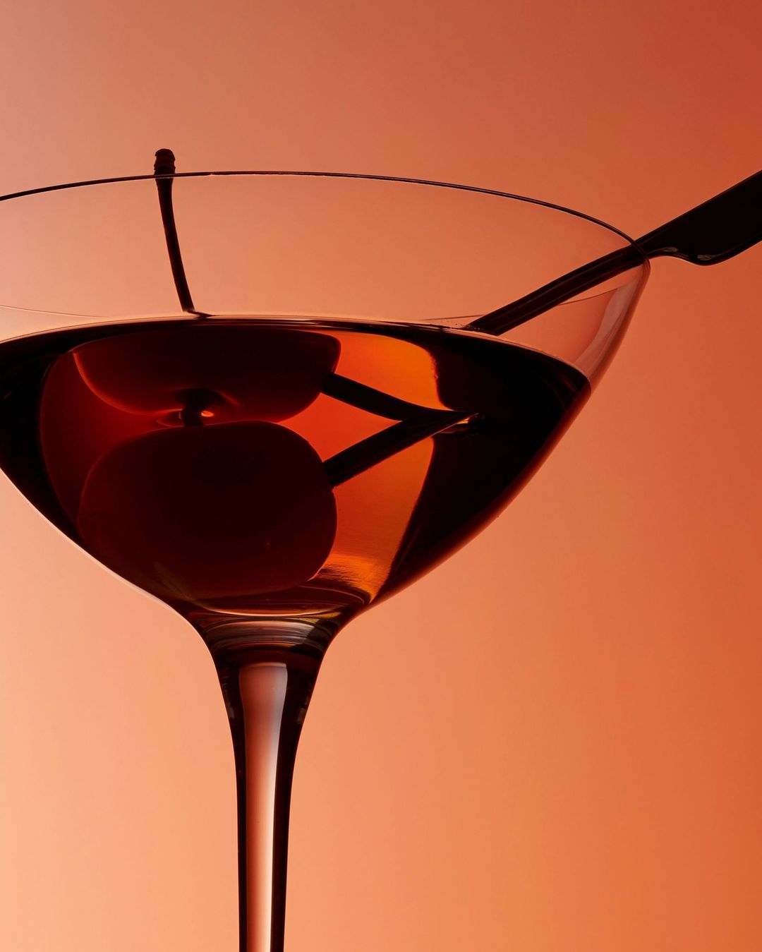Close-up studio shot of a stemmed cocktail glass with a dark amber liquid and two cherries, one skewered with a metal cocktail pick.