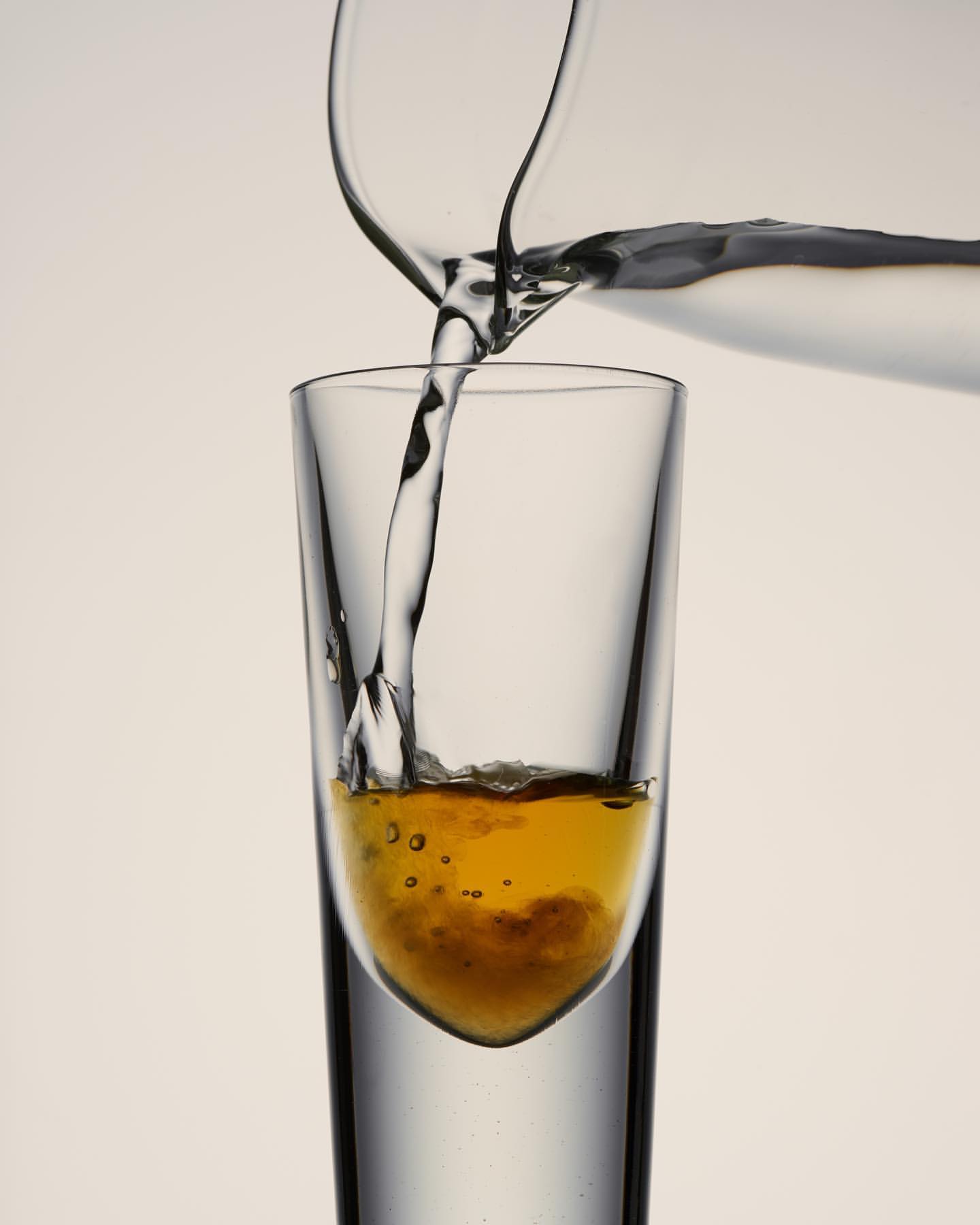 Clear pitcher pours liquid into a tall, slender glass. The liquid in the glass is amber with bubbles, against a plain background.
