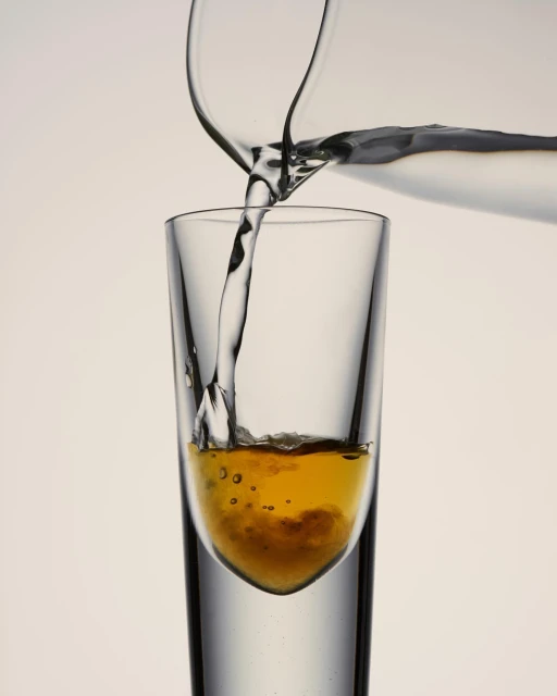 Clear pitcher pours liquid into a tall, slender glass. The liquid in the glass is amber with bubbles, against a plain background.