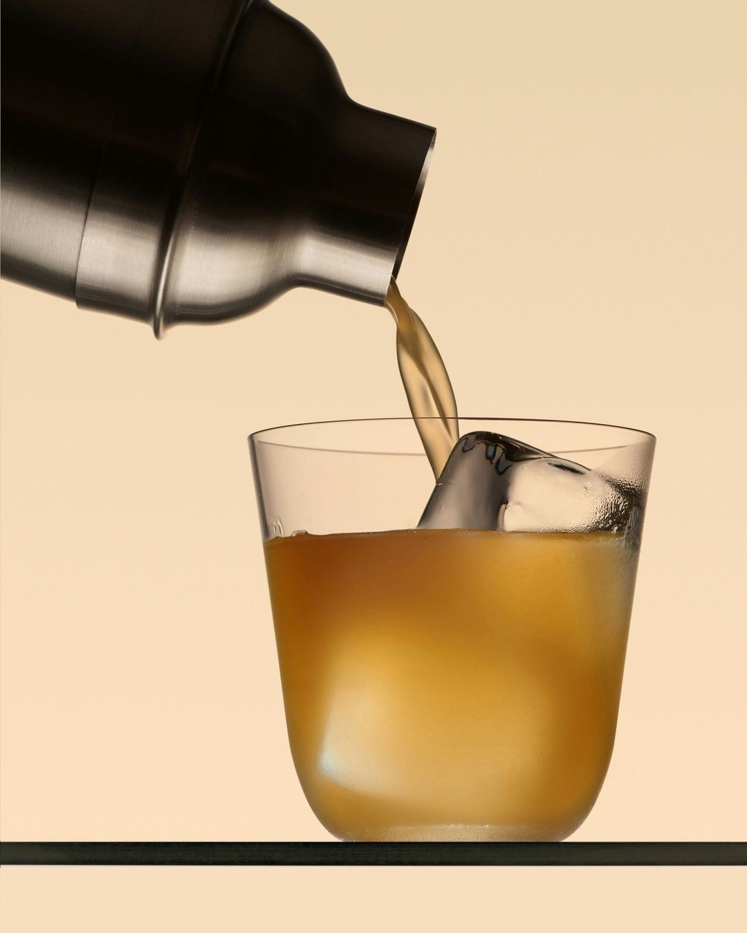 Drink being poured from a metal cocktail shaker into a clear glass with ice against a gradient light-yellow background.